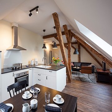 Esszimmer mit weißem Küchendesign und schwarzer Tischplatte. Im Hintergrund ein Wohnzimmer mit Holzbalken und schwarzem Sofa