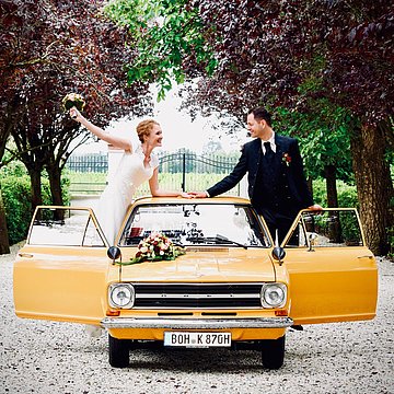 Heiratendendes Ehepaar stehen auf einem gelben Auto, auf welchem ein Blumenstrauß liegt und lächeln sich gegenseitig an.